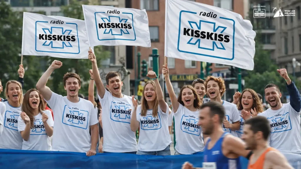 Milano Marathon 2026: Tifosi di Radio Kiss Kiss applaudono e sventolano tre bandiere con il logo dell’emittente durante la Milano Marathon, mentre alcuni corridori passano in primo piano. In alto a destra compaiono i loghi di Radio Kiss Kiss e Wizz Air Milano Marathon.