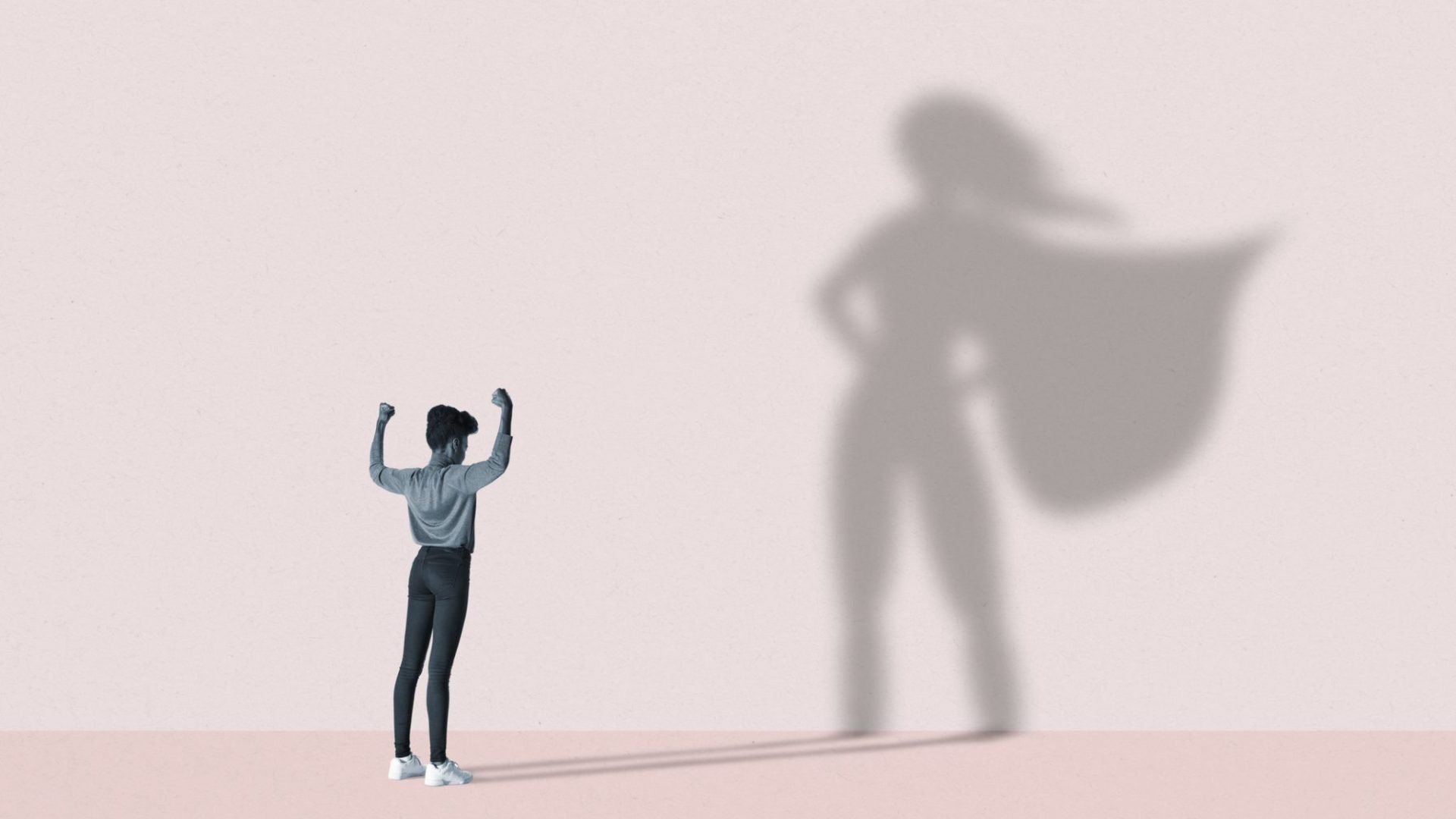 Young woman flexing muscles in front of large superhero shadow on pink background