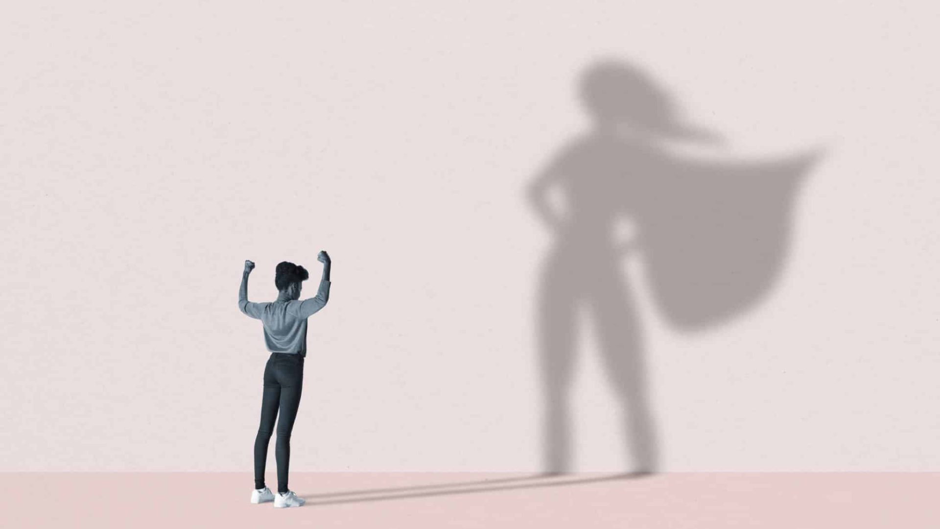 Young woman flexing muscles in front of large superhero shadow on pink background