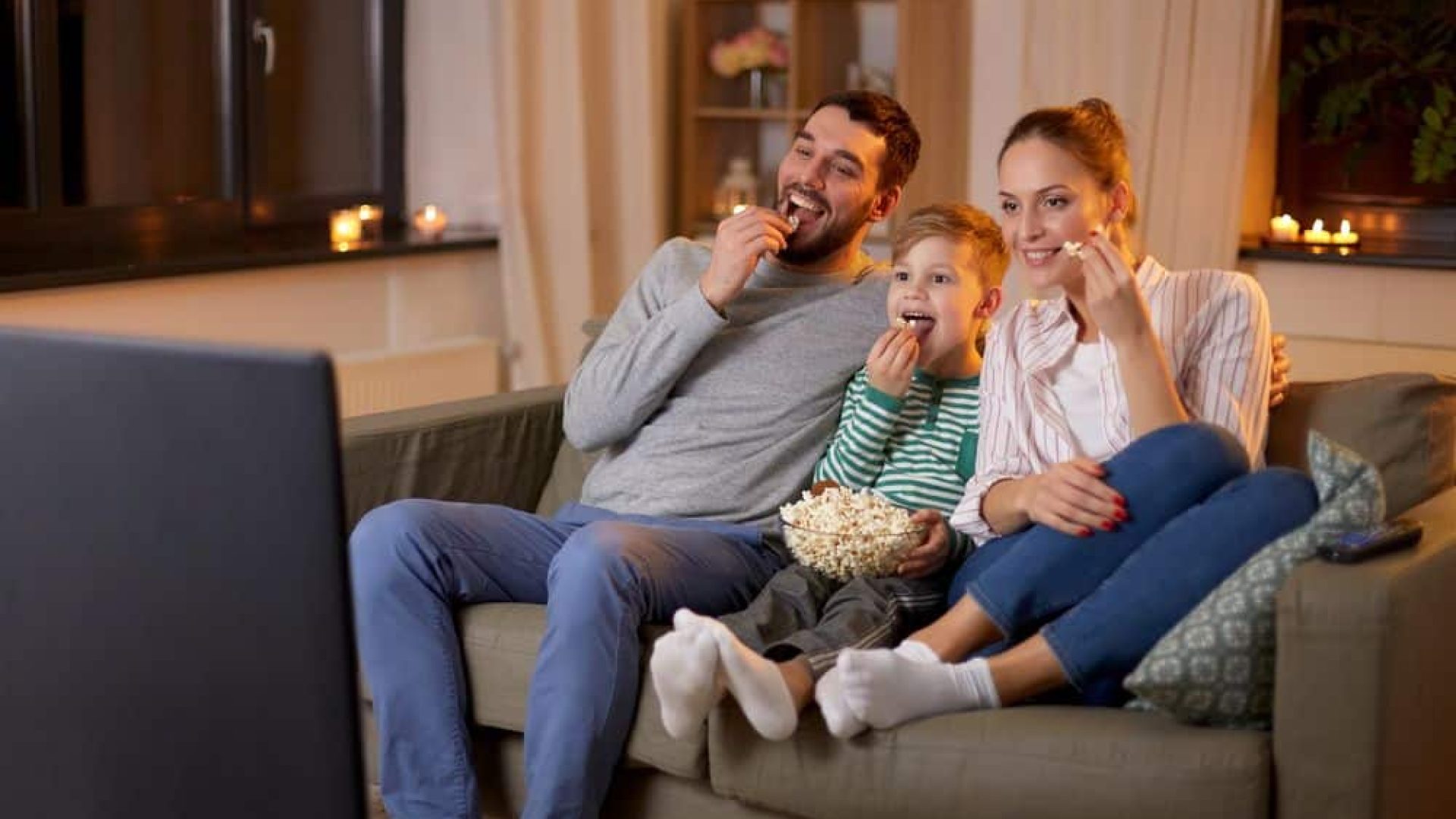 concetto di famiglia, tempo libero e persone - padre sorridente felice, madre e figlio piccolo guardando la tv e mangiando popcorn a casa di notte
